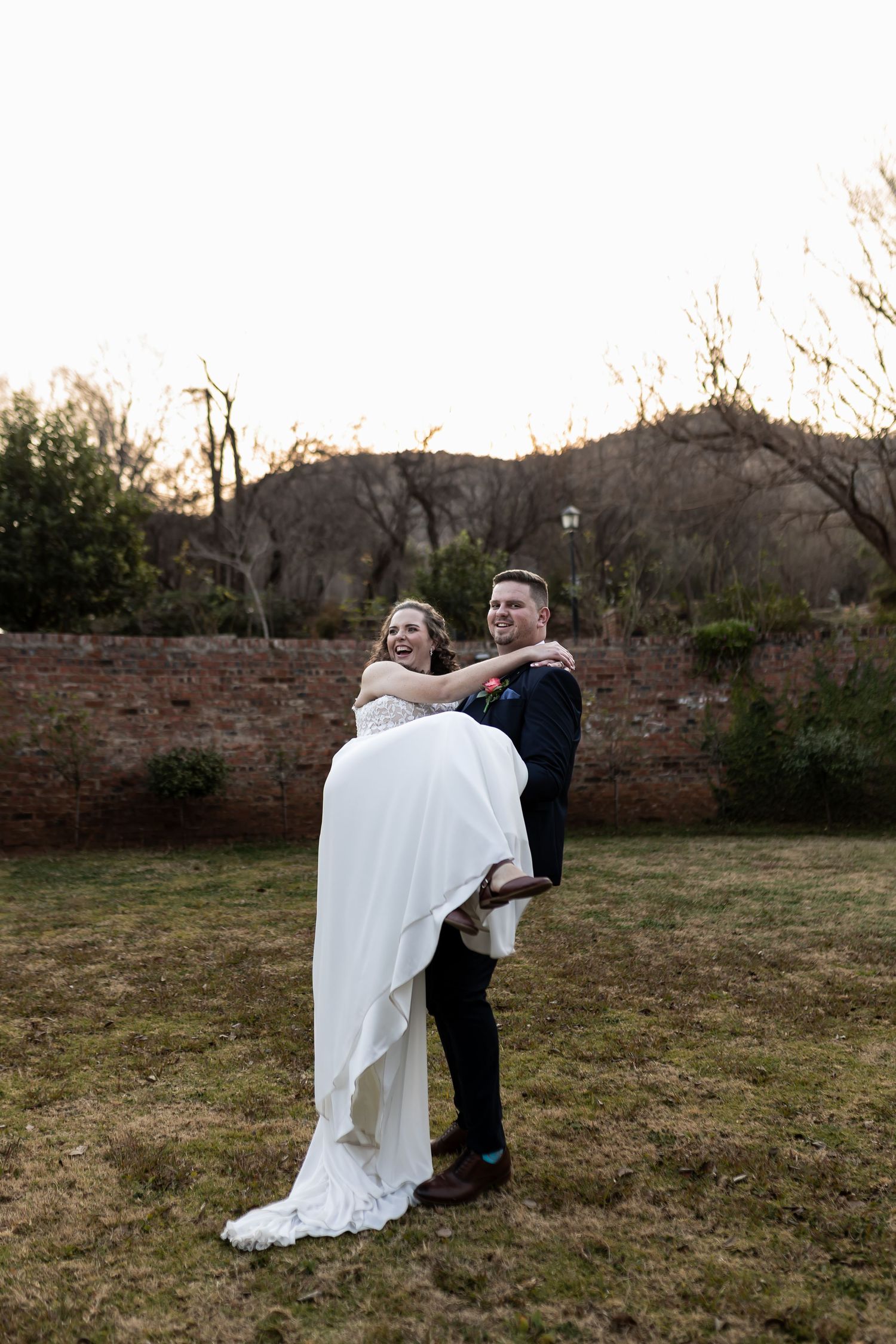 A romantic wedding couple embraces at dusk in front of a rustic stone wall as the groom carries his bride in her flowing gown.