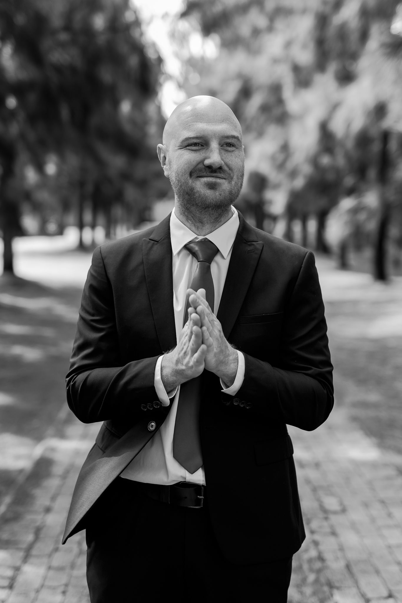 Man in dark suit poses with clasped hands on brick pathway in black and white photo.
