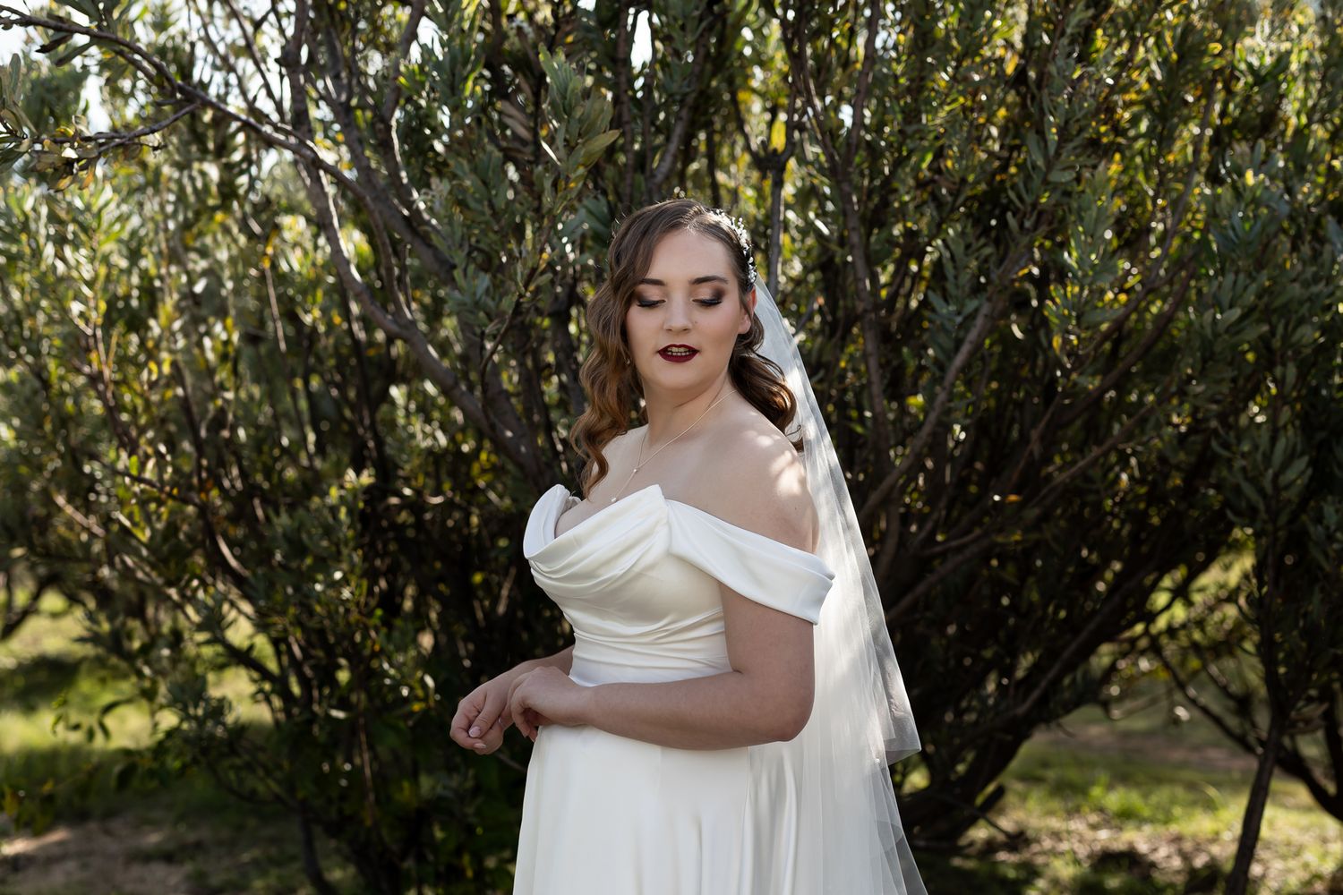 Bride in classic white off-shoulder wedding gown poses beside green foliage.