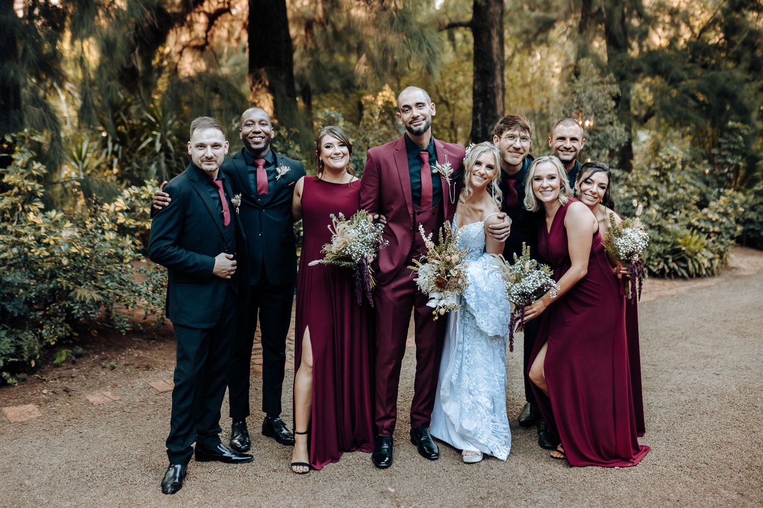 Wedding party poses in burgundy and white attire on garden path.