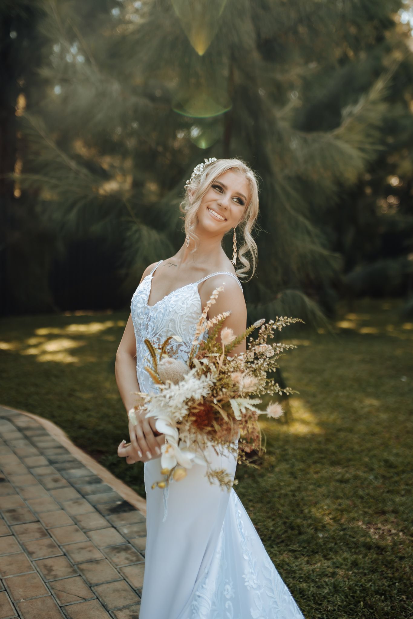 Bride in lace dress holds dried flower bouquet while posing on brick pathway in garden setting.