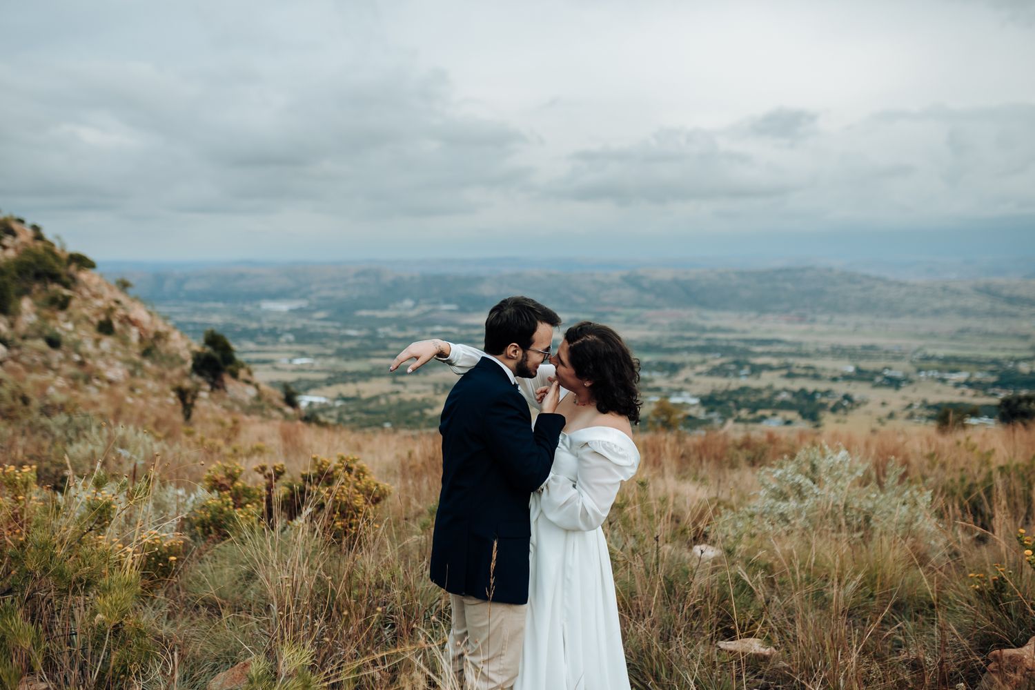 A romantic wedding portrait of a couple against a dramatic mountain landscape and cloudy sky.
