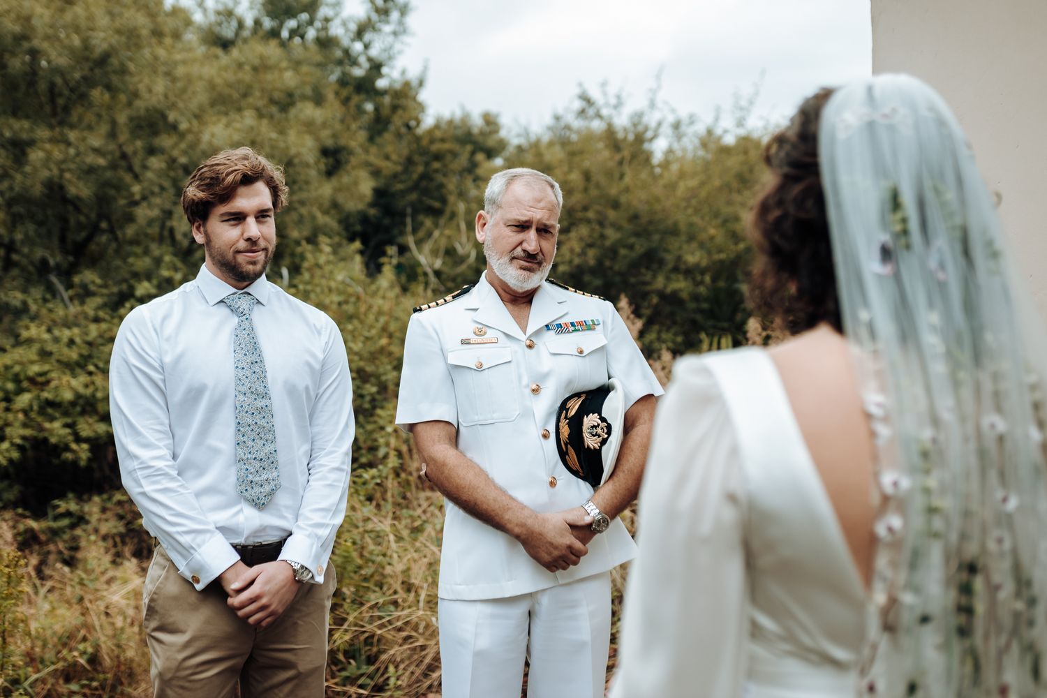 Outdoor ceremony scene with officiant in white uniform and attendees in formal wear.