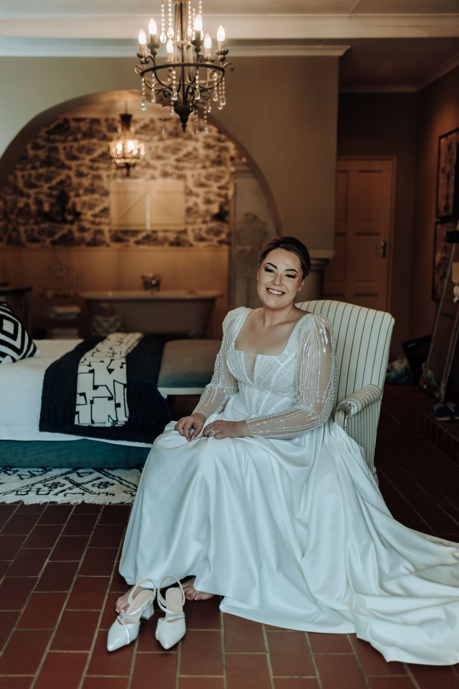 Bride in elegant long-sleeved wedding dress sitting in vintage room with stone wall and chandelier.