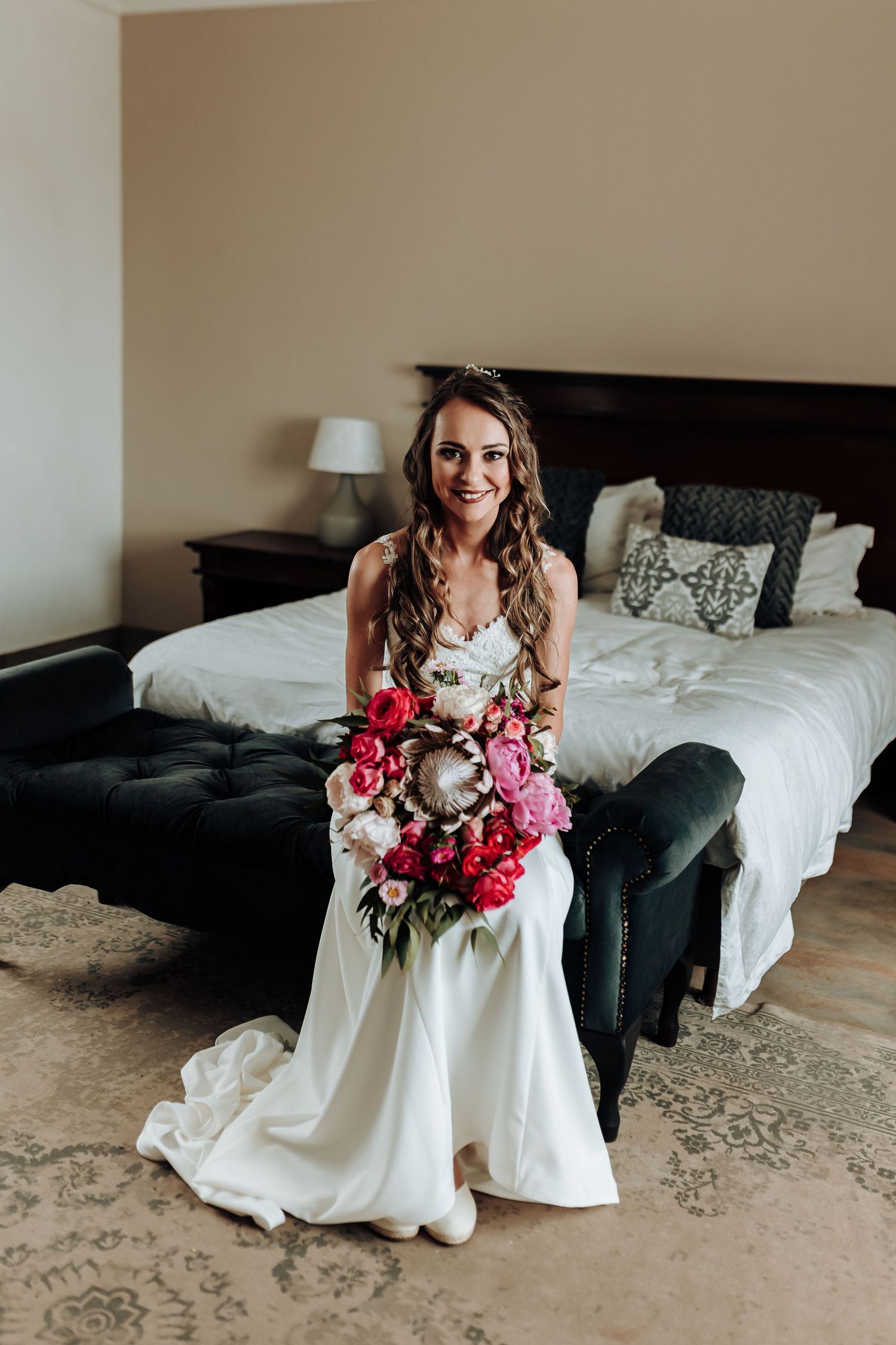 Bride in white gown sits on dark couch holding vibrant pink and white bouquet.
