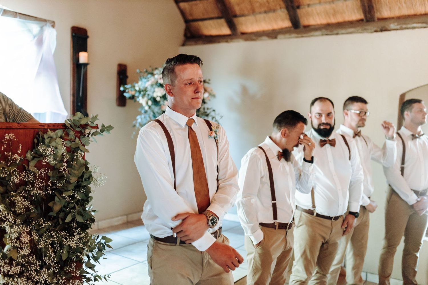 Groomsmen in white shirts and suspenders stand in formation at a rustic wedding venue.