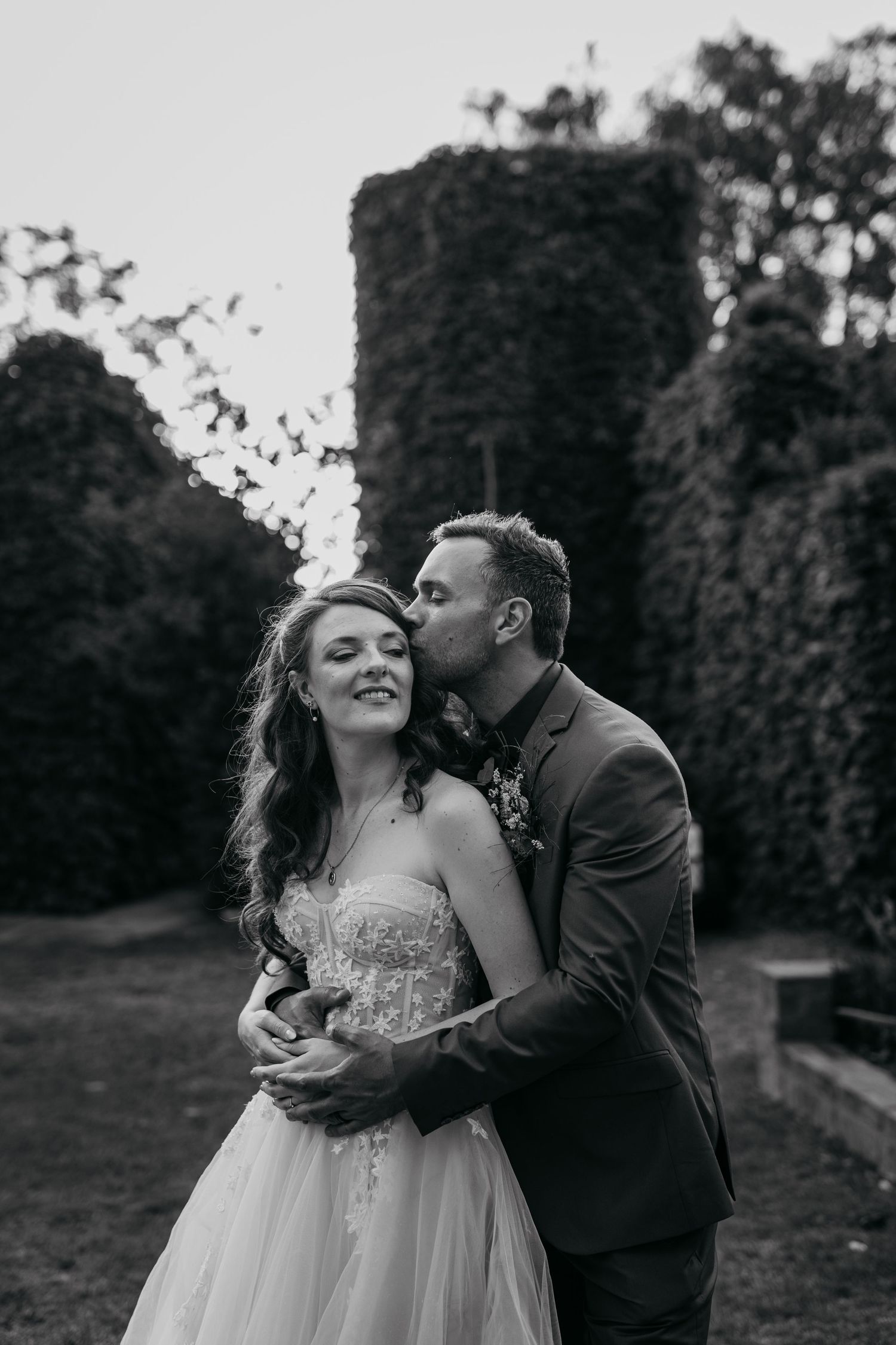 A romantic black and white wedding photo of a couple embracing in front of ivy-covered walls at sunset.