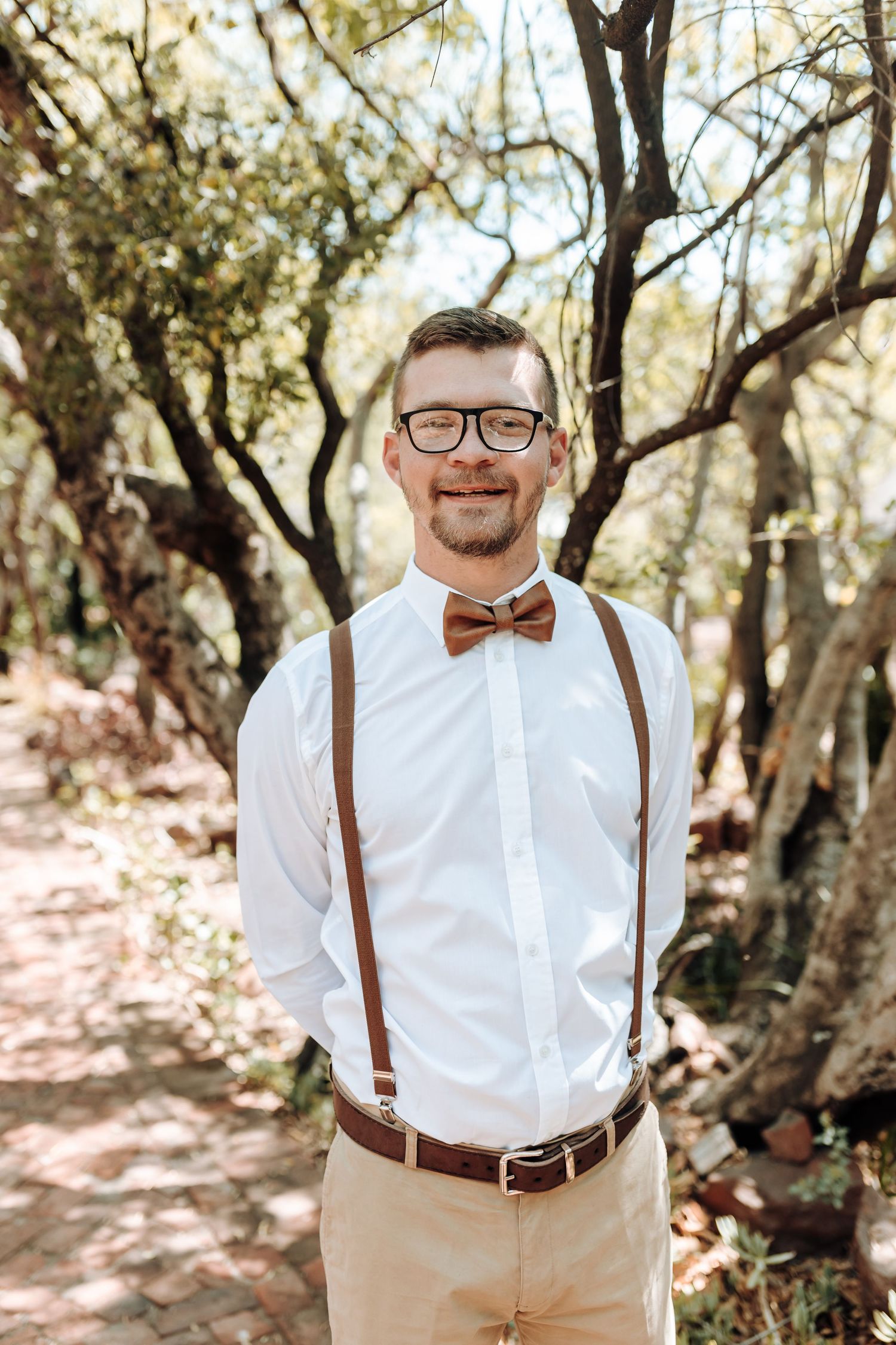 Groomsman in brown bow tie and suspenders smiles while standing among trees at outdoor wedding.