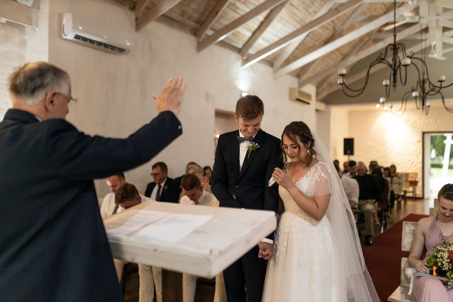 A wedding officiant raises his hand during a ceremony in a light-filled venue with wooden beams.
