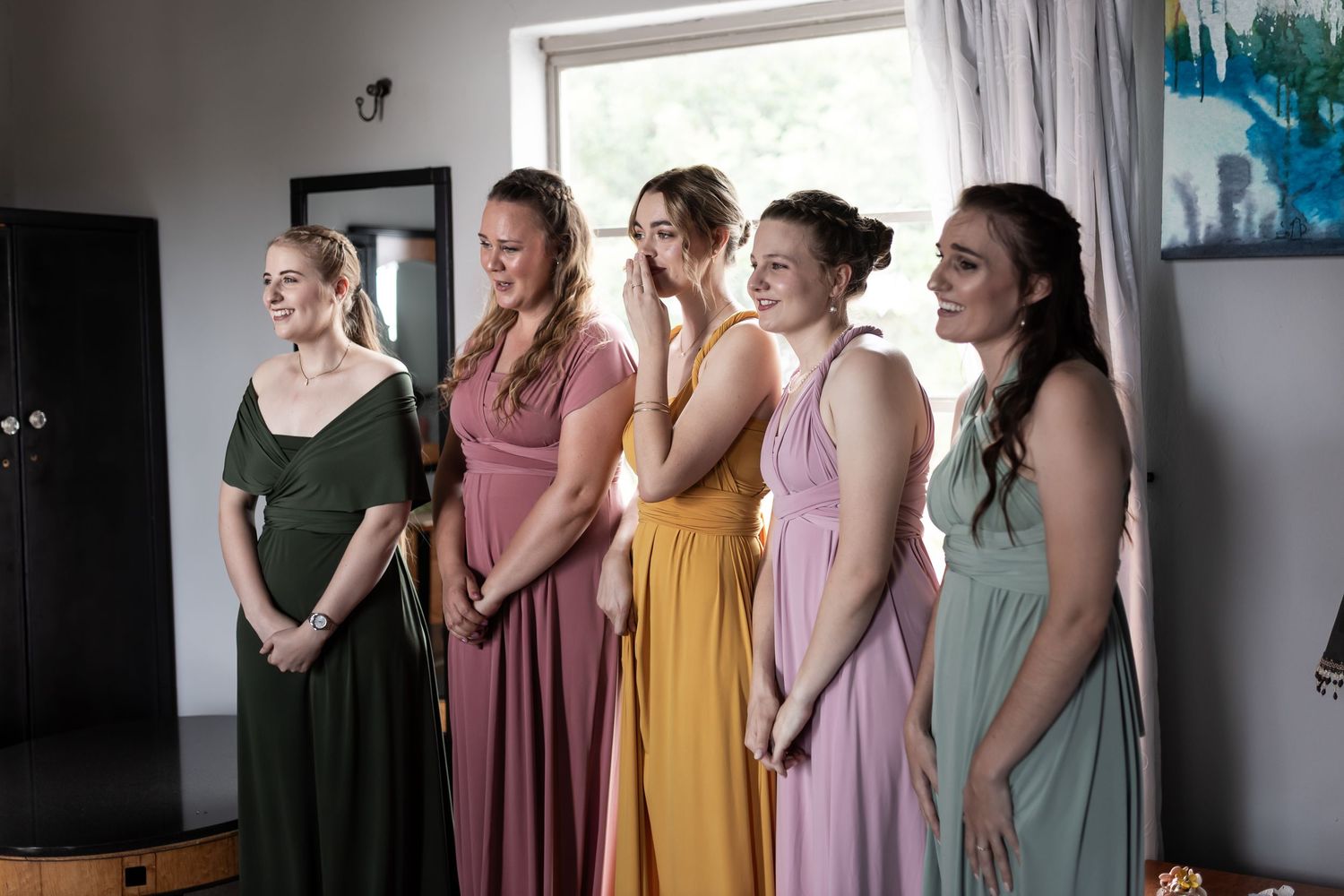 A group of bridesmaids in different colored dresses stand together in a room with blue wall art.
