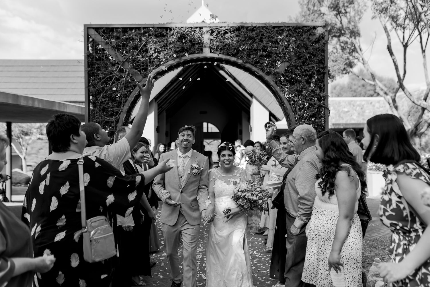 Wedding guests celebrating and throwing confetti as newlyweds exit through decorative archway at outdoor ceremony.