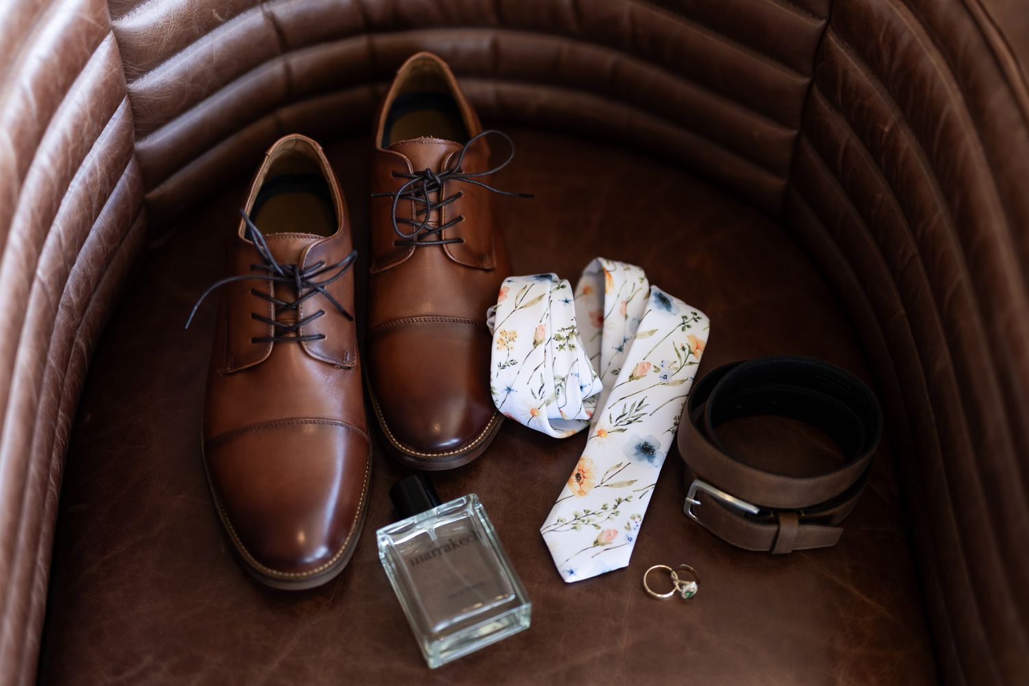 Wedding accessories including brown leather shoes, socks, cologne and belt arranged on a brown leather chair.