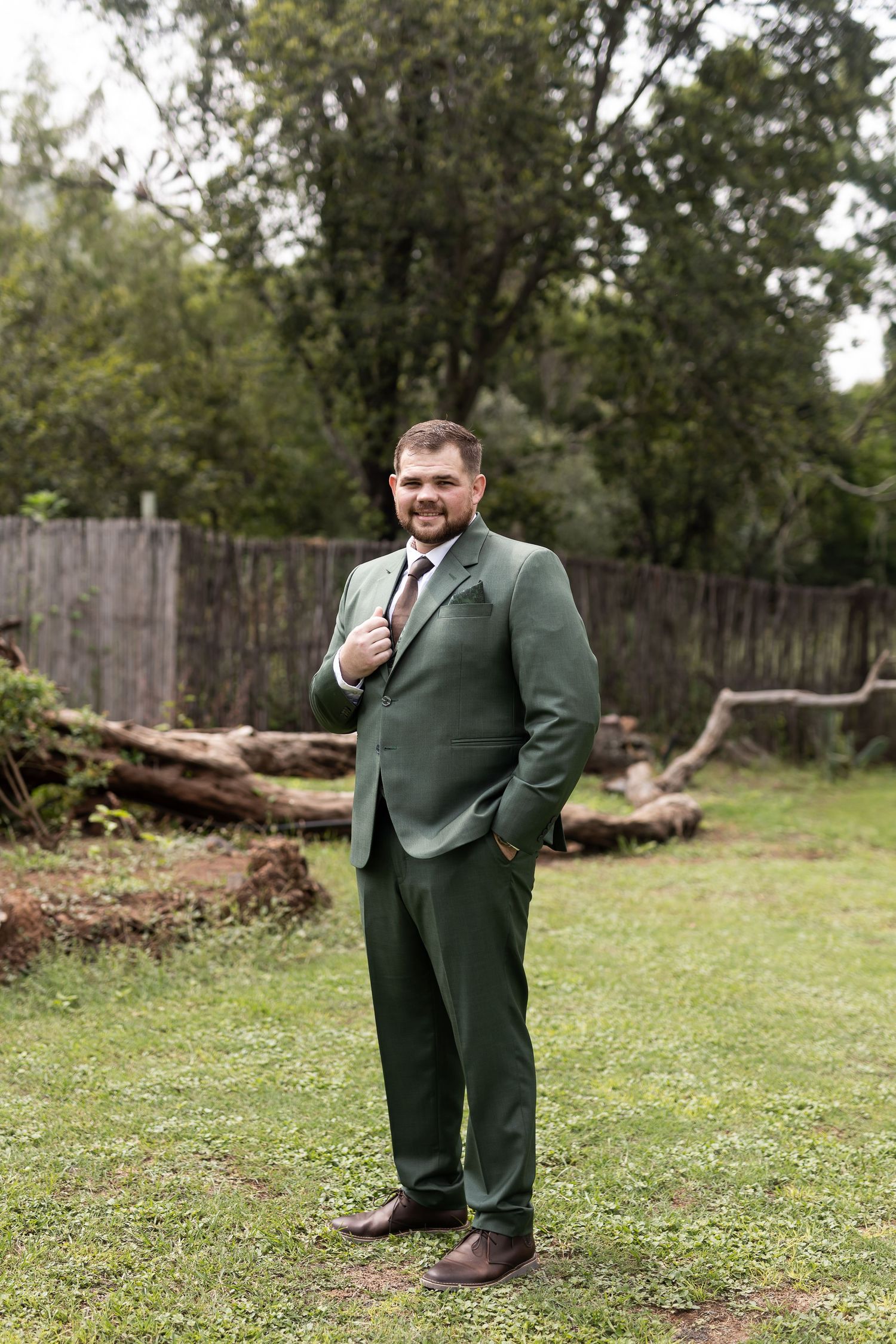 A person in a forest green suit poses confidently in a backyard garden setting with a wooden fence.