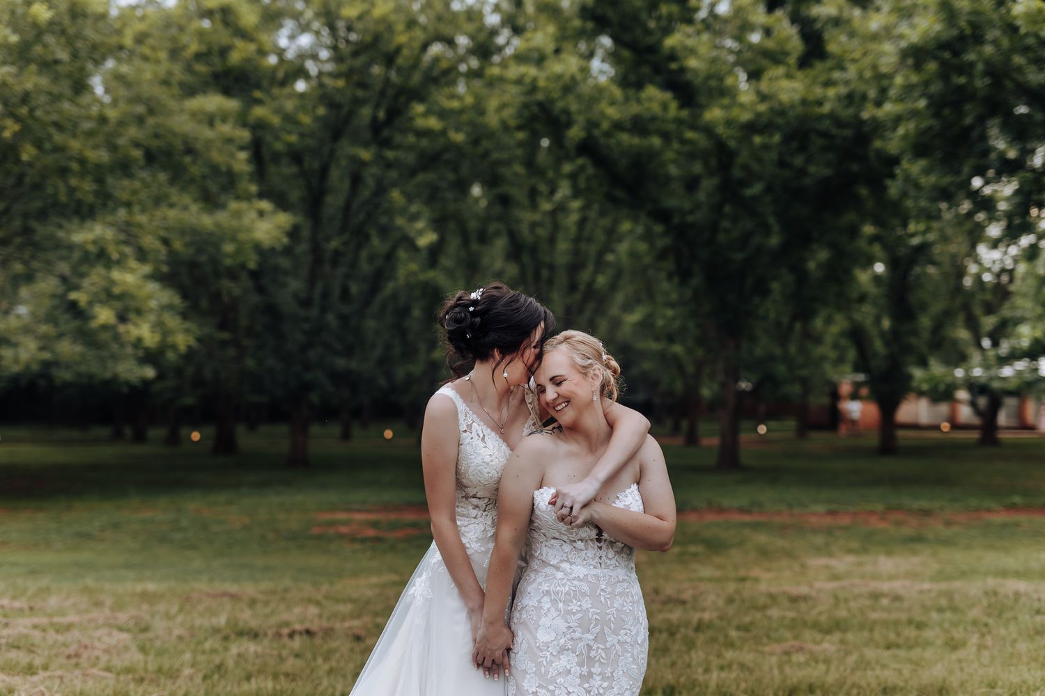 Two brides in elegant white dresses share a joyful laugh together in a park setting.
