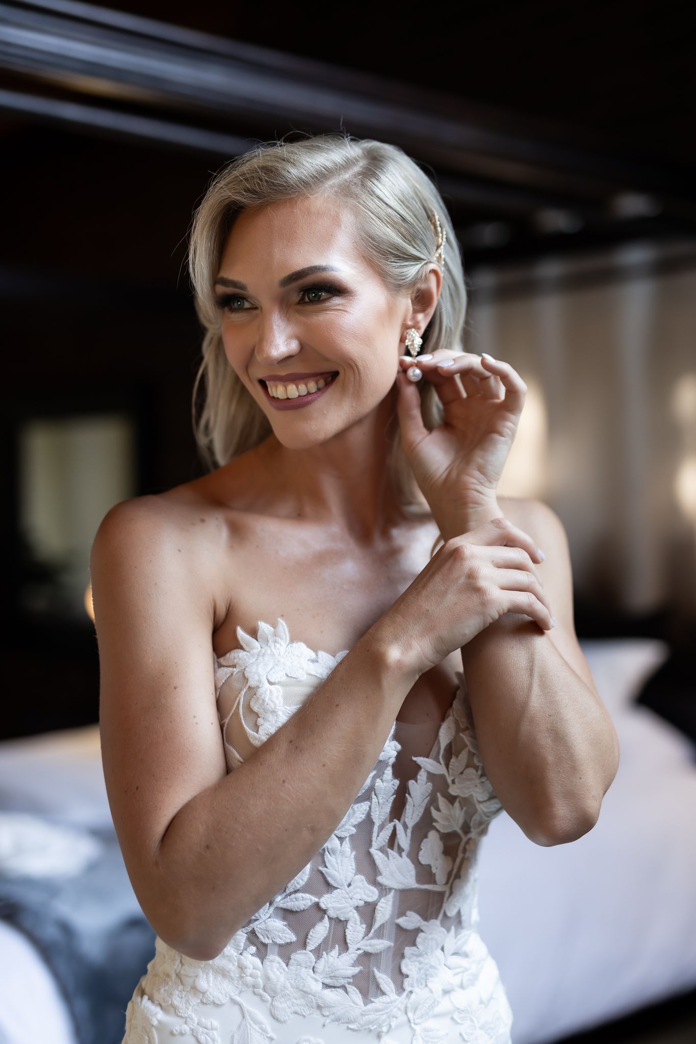 Close-up of bride in strapless lace gown smiling while putting on jewelry.