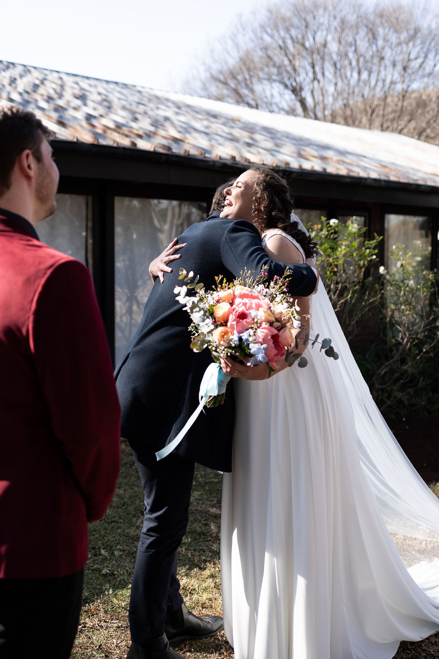 A wedding celebration showing an embrace during an outdoor ceremony, with a beautiful bouquet of pink and white flowers.