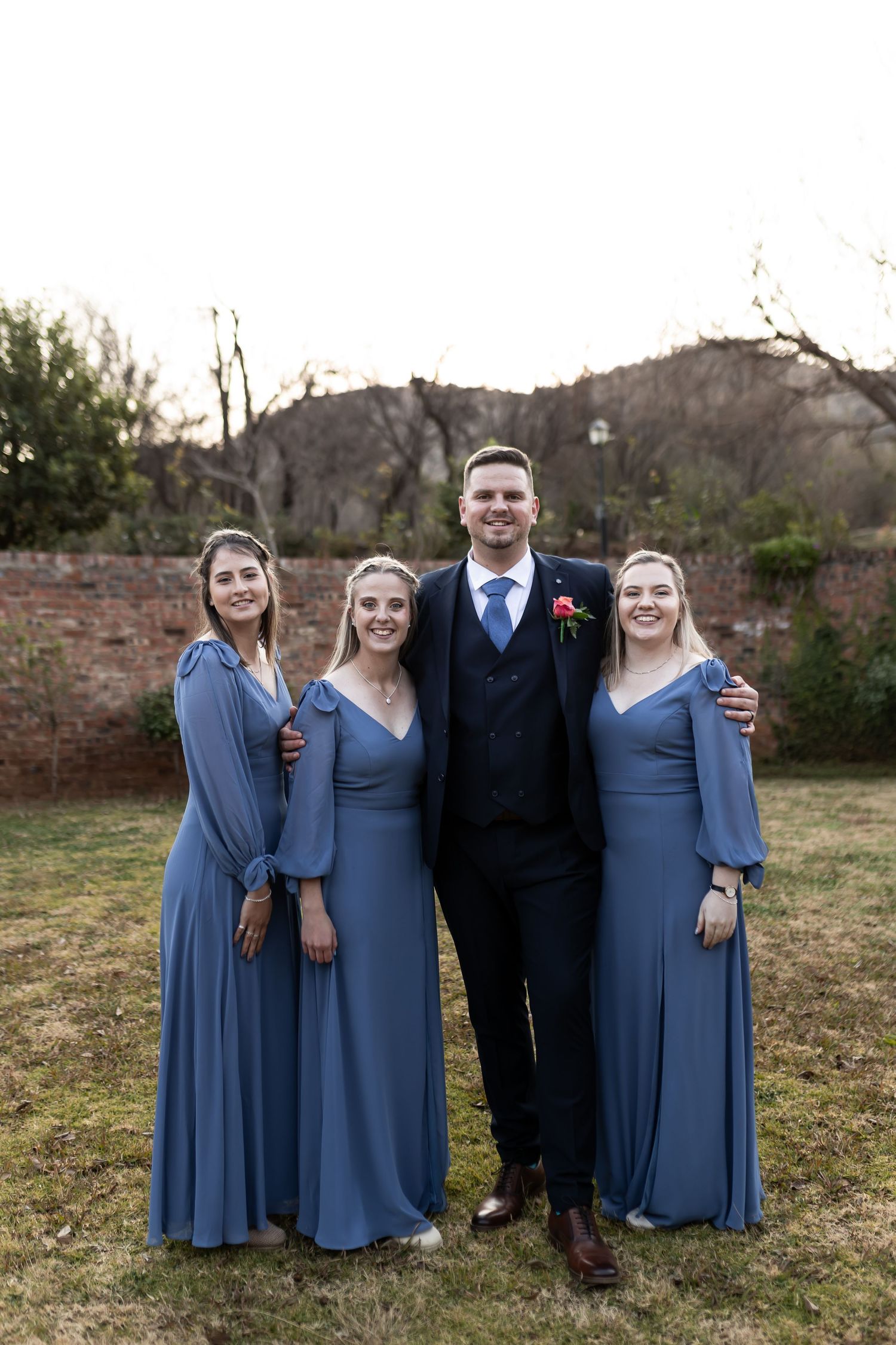 Wedding party group poses outdoors in front of stone wall with person in navy suit and three in dusty blue gowns.