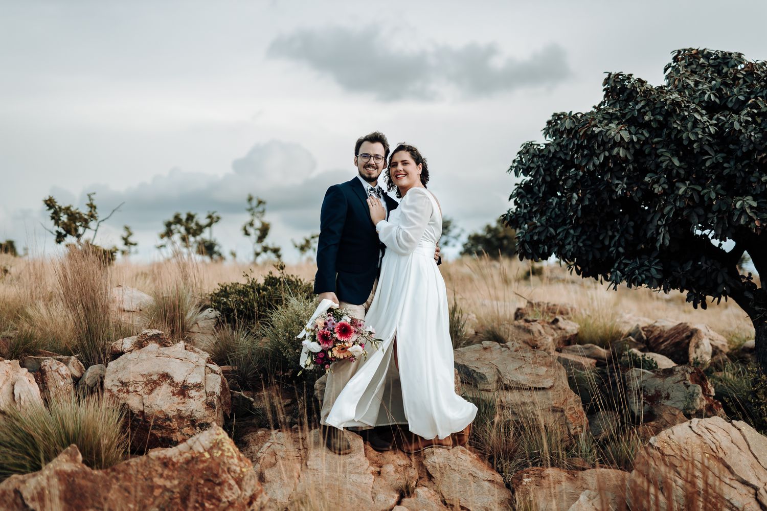 A wedding couple poses on rocky terrain with cloudy skies and mountains in the background.