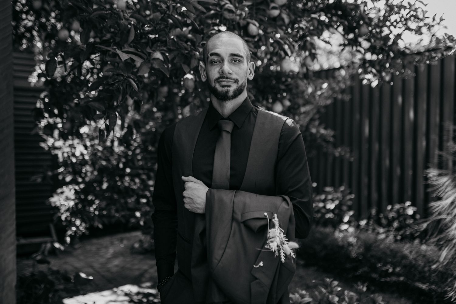 A groom in a black suit poses for a portrait in a garden setting.