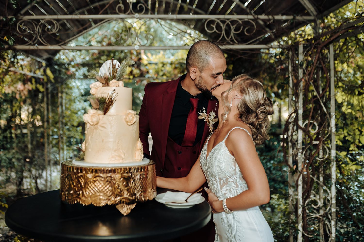 Couple shares a romantic kiss next to their elegant gold and white wedding cake under a decorative gazebo.