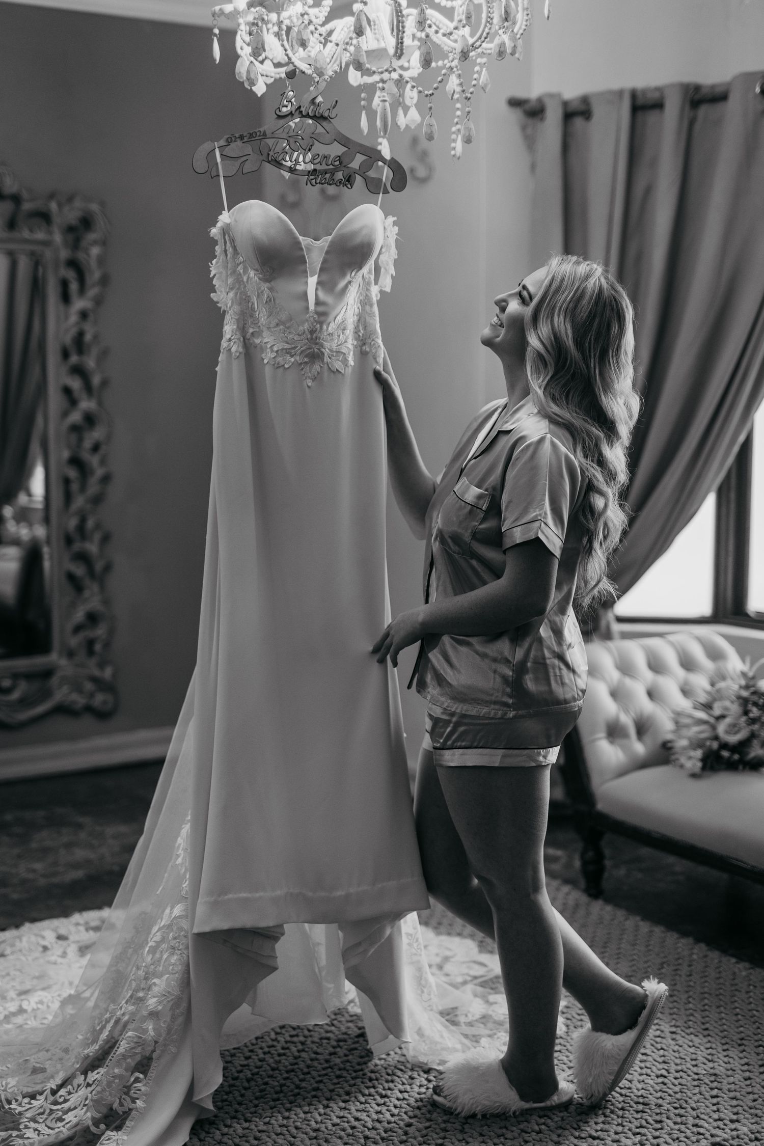 Black and white photo of wedding dress suspended from chandelier being admired.