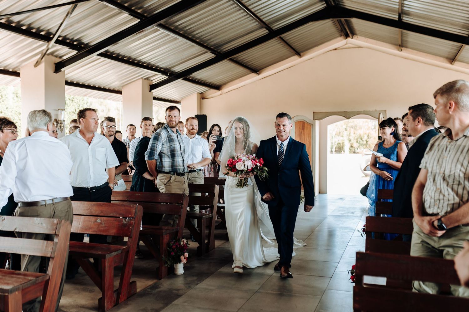 Wedding guests line wooden pews in a covered venue as a ceremony procession walks down the aisle.