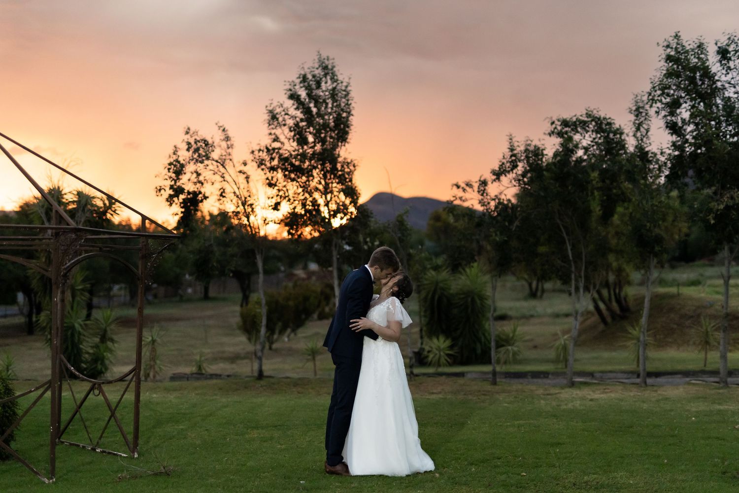 Newlyweds embrace during sunset with mountains and rustic venue in the background.