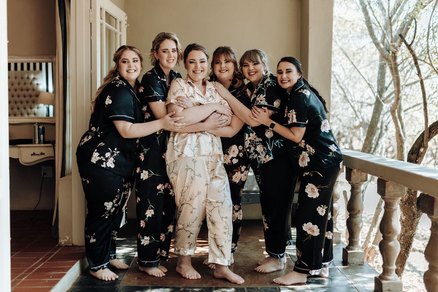 Wedding party in coordinated floral robes hugging together on a porch.