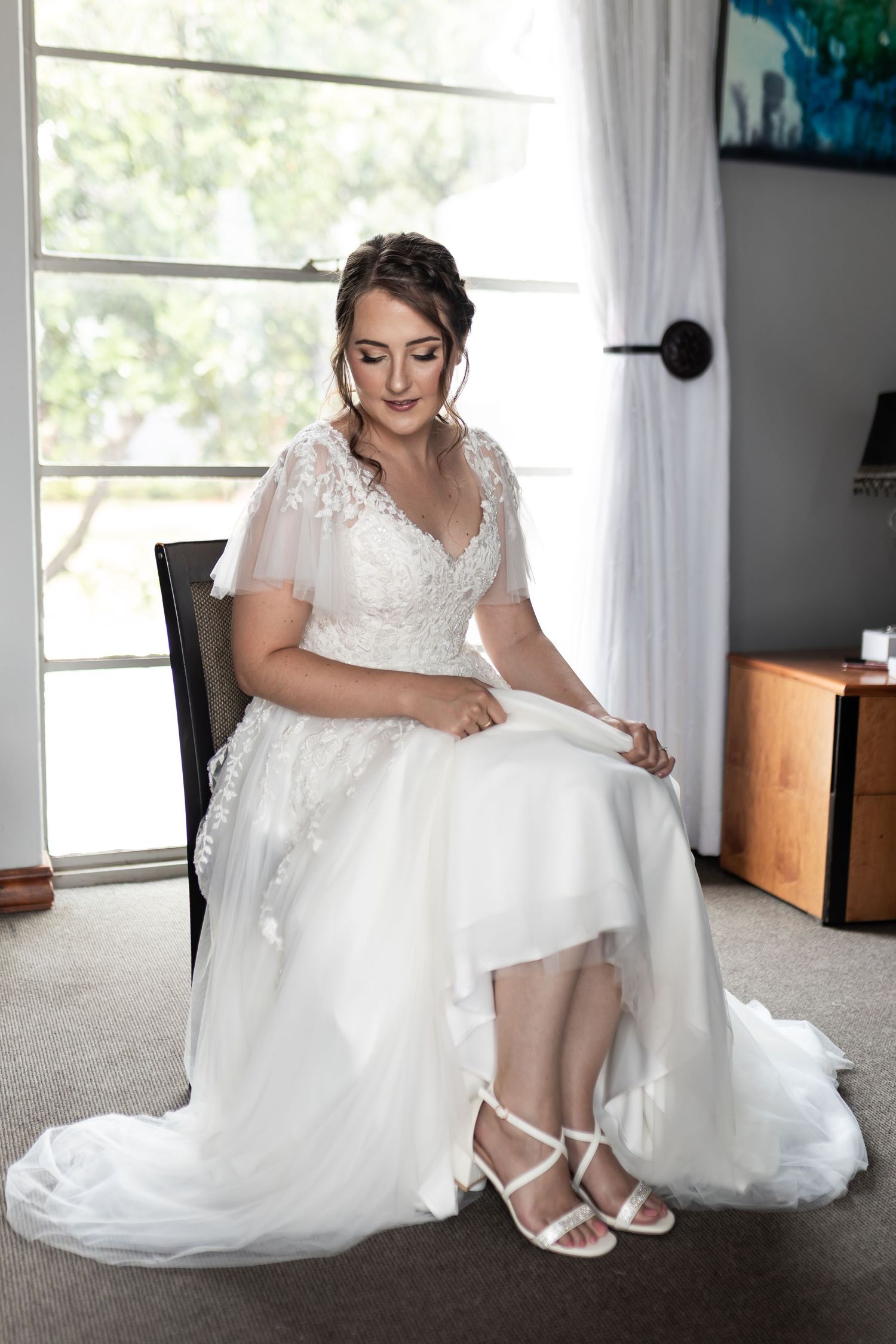 A person in a white lace wedding gown sits elegantly in a chair by a window.