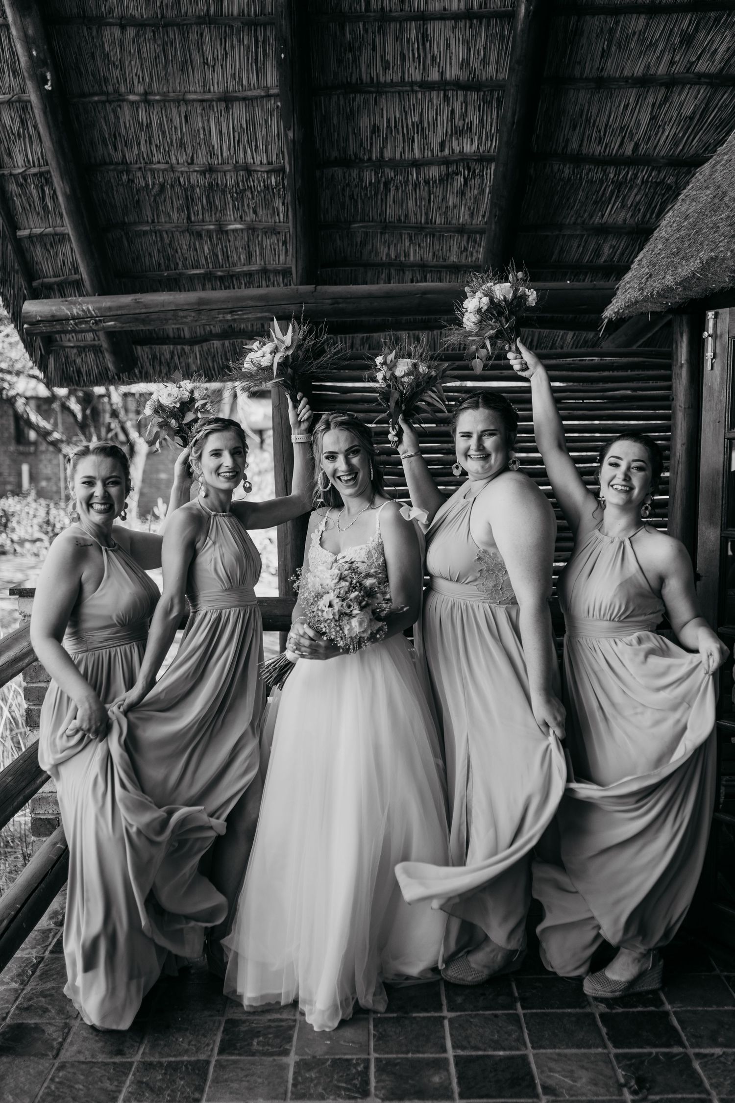 Black and white photo of bridal party celebrating with raised bouquets under thatched roof.
