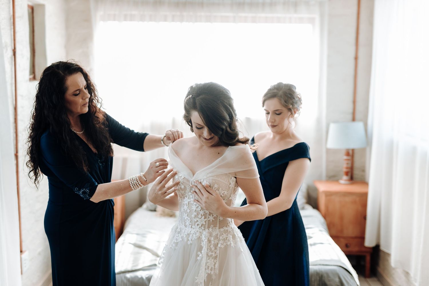 Women in navy dresses helping adjust a bride's wedding gown in a bedroom setting.