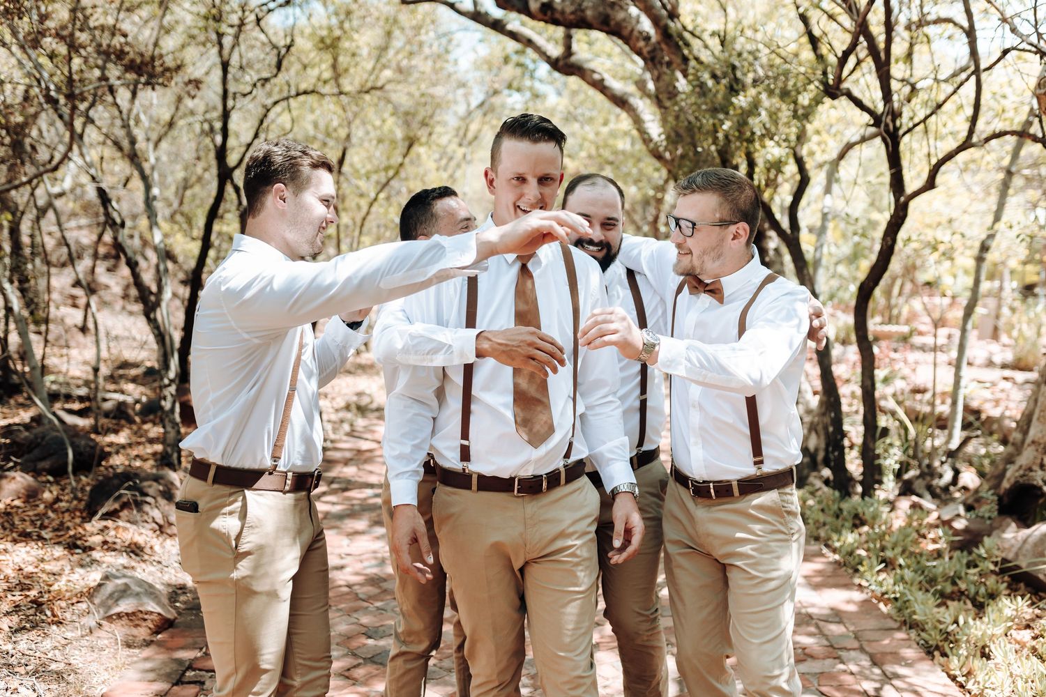 Wedding party in coordinated attire toasting drinks together on a wooded path.