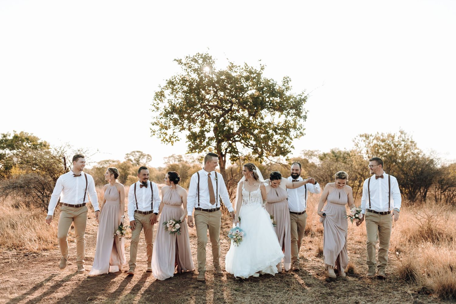 Wedding party in beige and white attire poses together under a lone tree.
