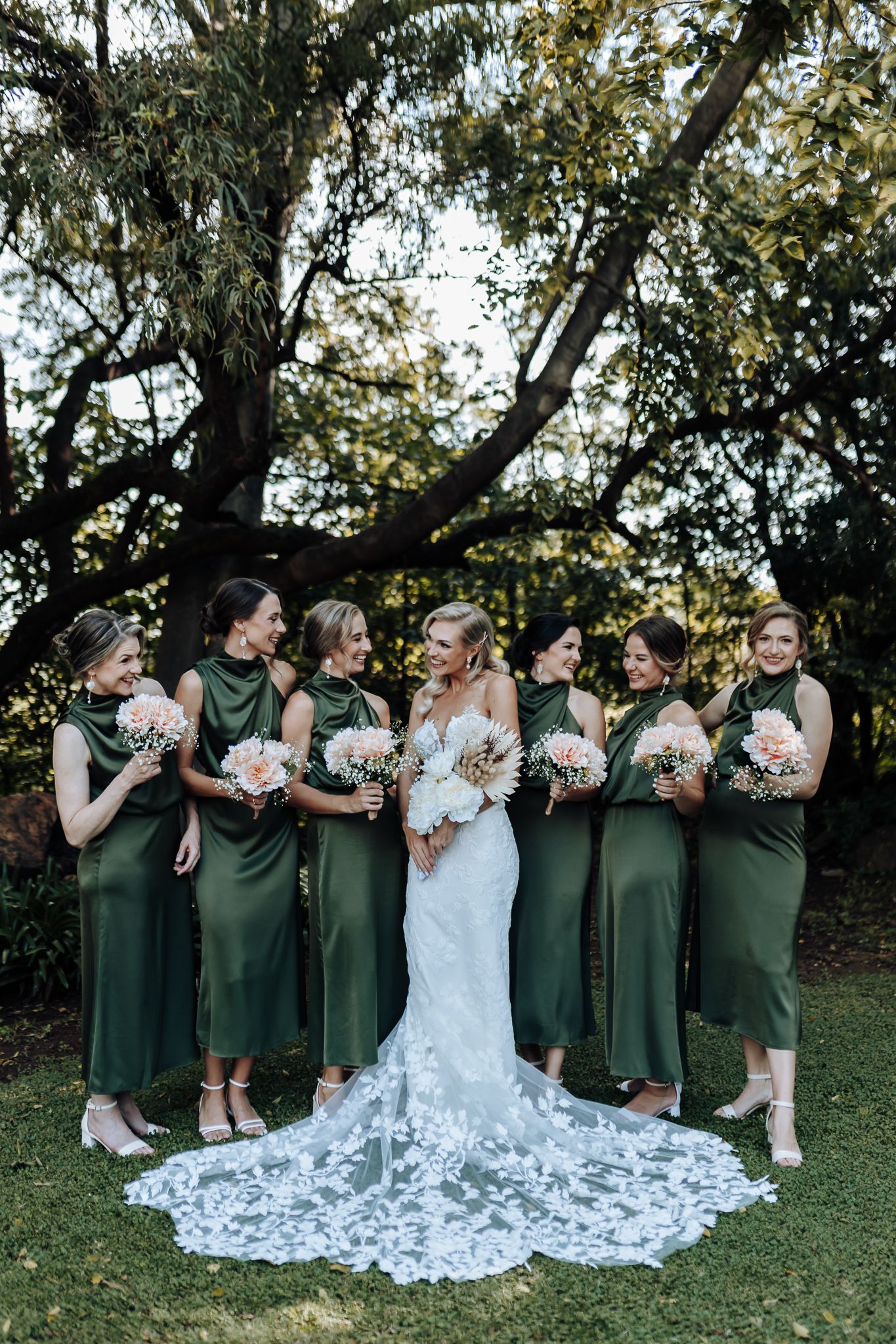 Bridal party in green dresses shares candid moment outdoors under tree canopy.
