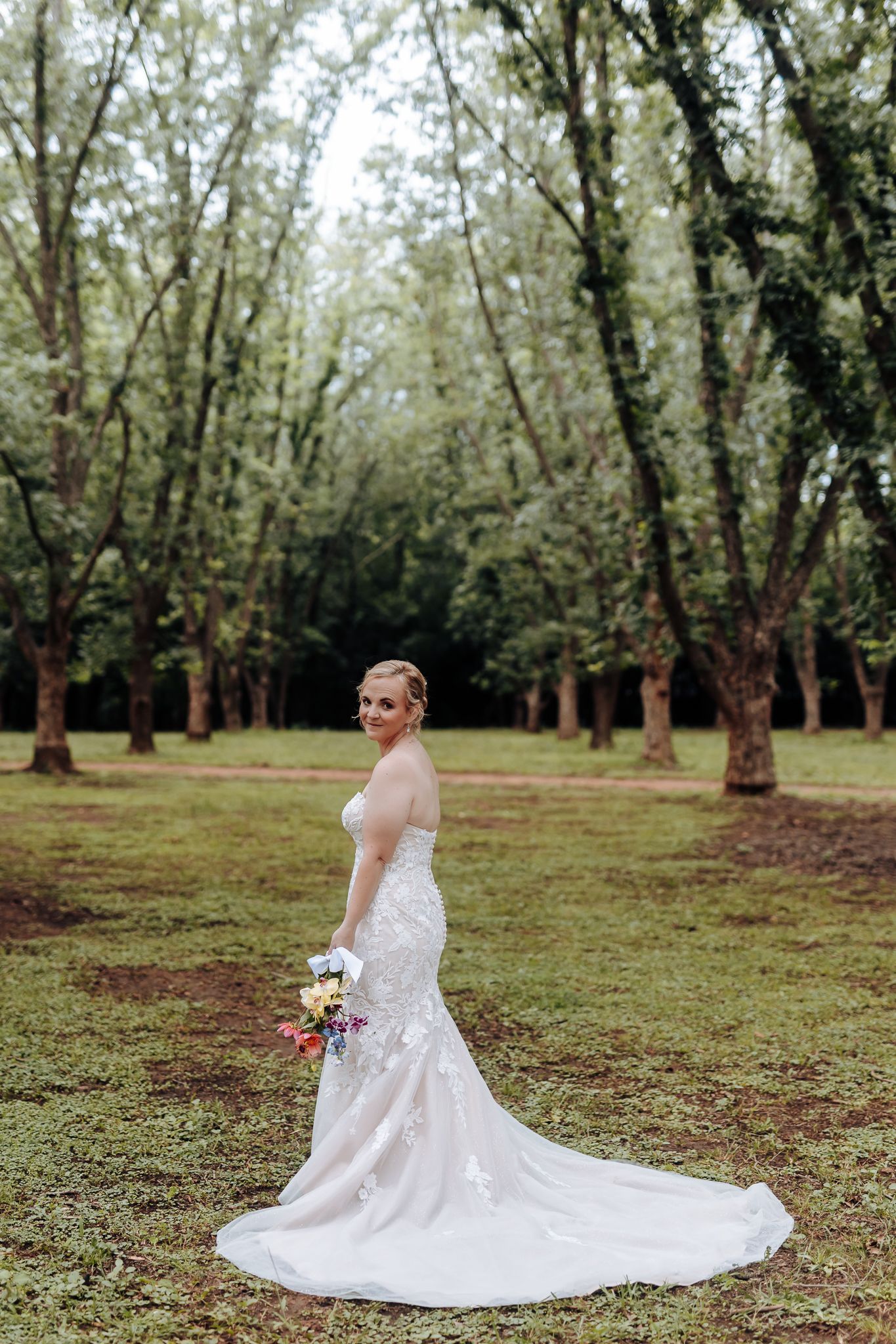 A stunning bride in a lace wedding gown poses gracefully in a wooded area holding a colorful bouquet.
