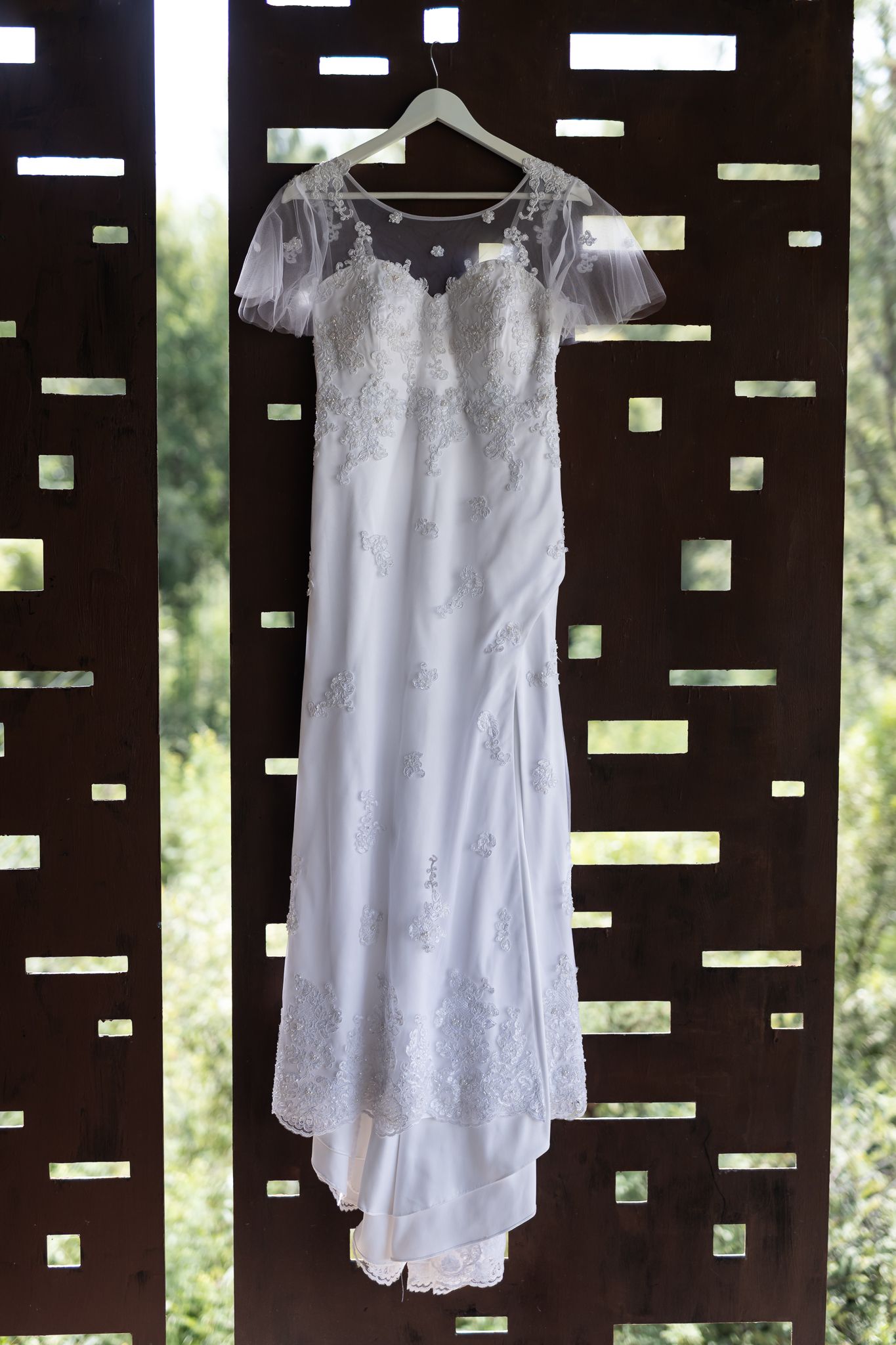 White lace wedding dress hanging on display against a dark wooden backdrop with geometric cutouts.