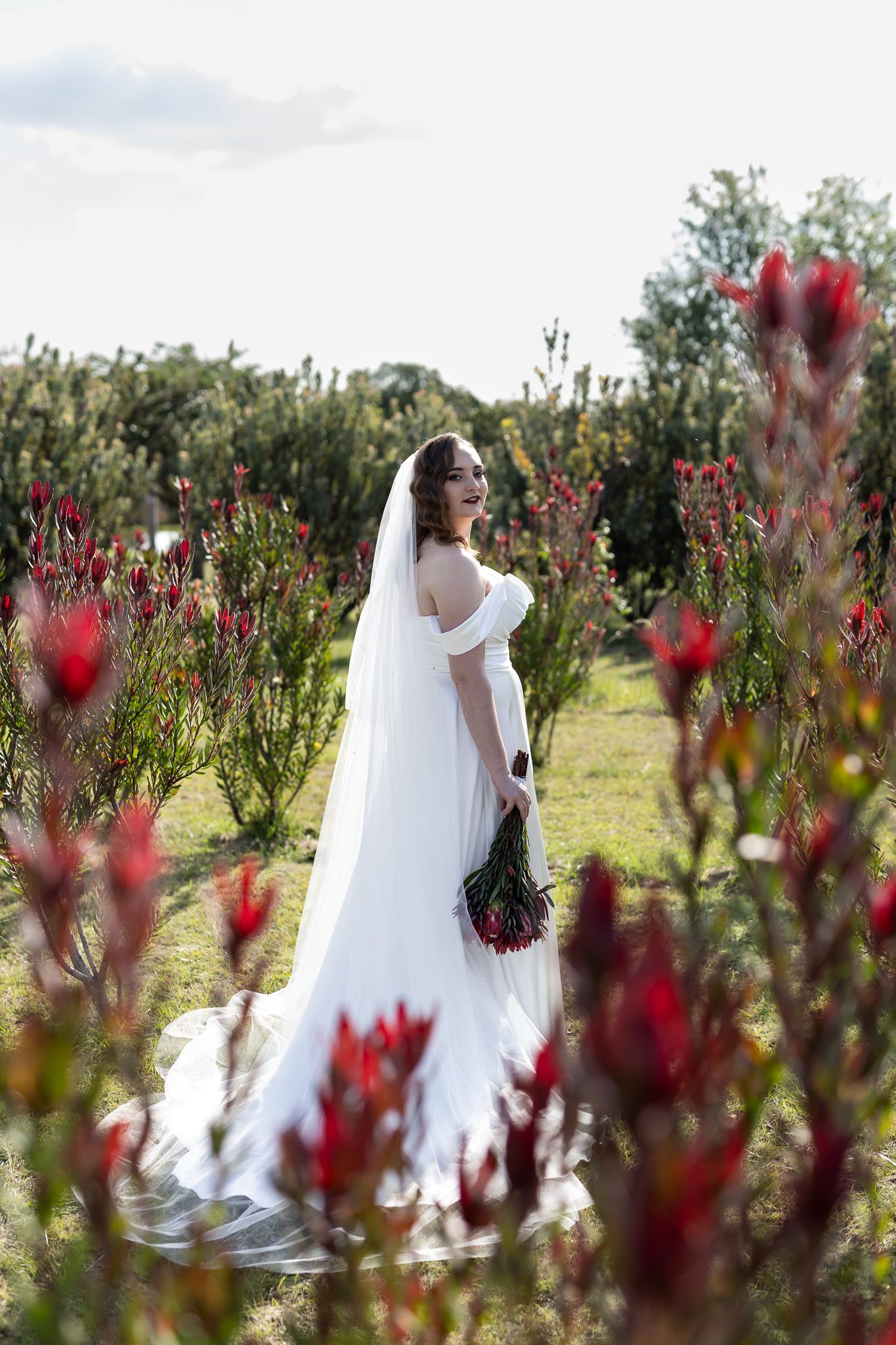 Bride in flowing white dress poses among red flowering plants in garden setting.