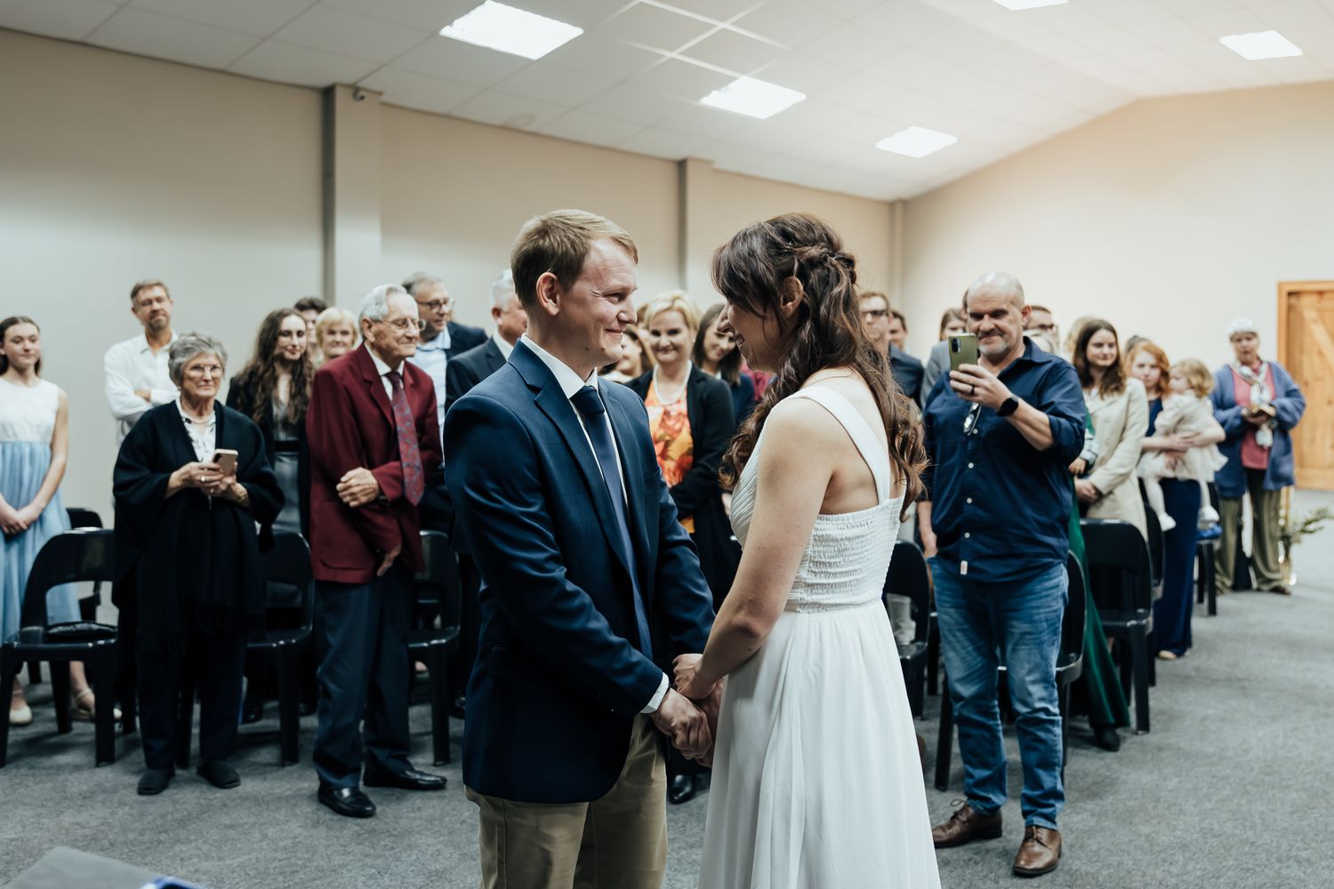 Wedding ceremony guests seated in rows at an indoor venue with fluorescent lighting.