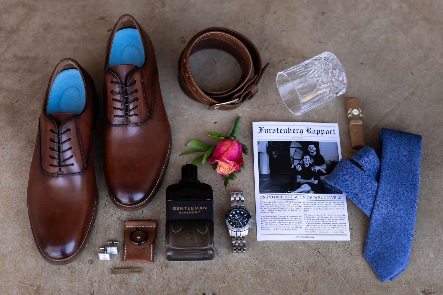 Brown leather dress shoes arranged with accessories including a blue tie, leather belt, watch, and perfume bottle.