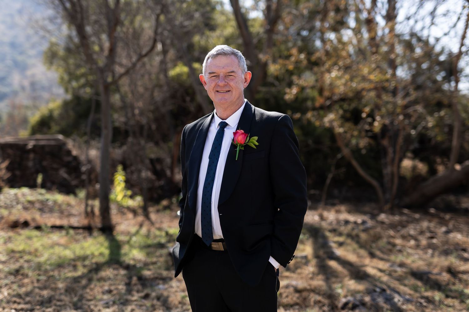 A person in a dark suit and tie with a red boutonniere stands outdoors in natural lighting.