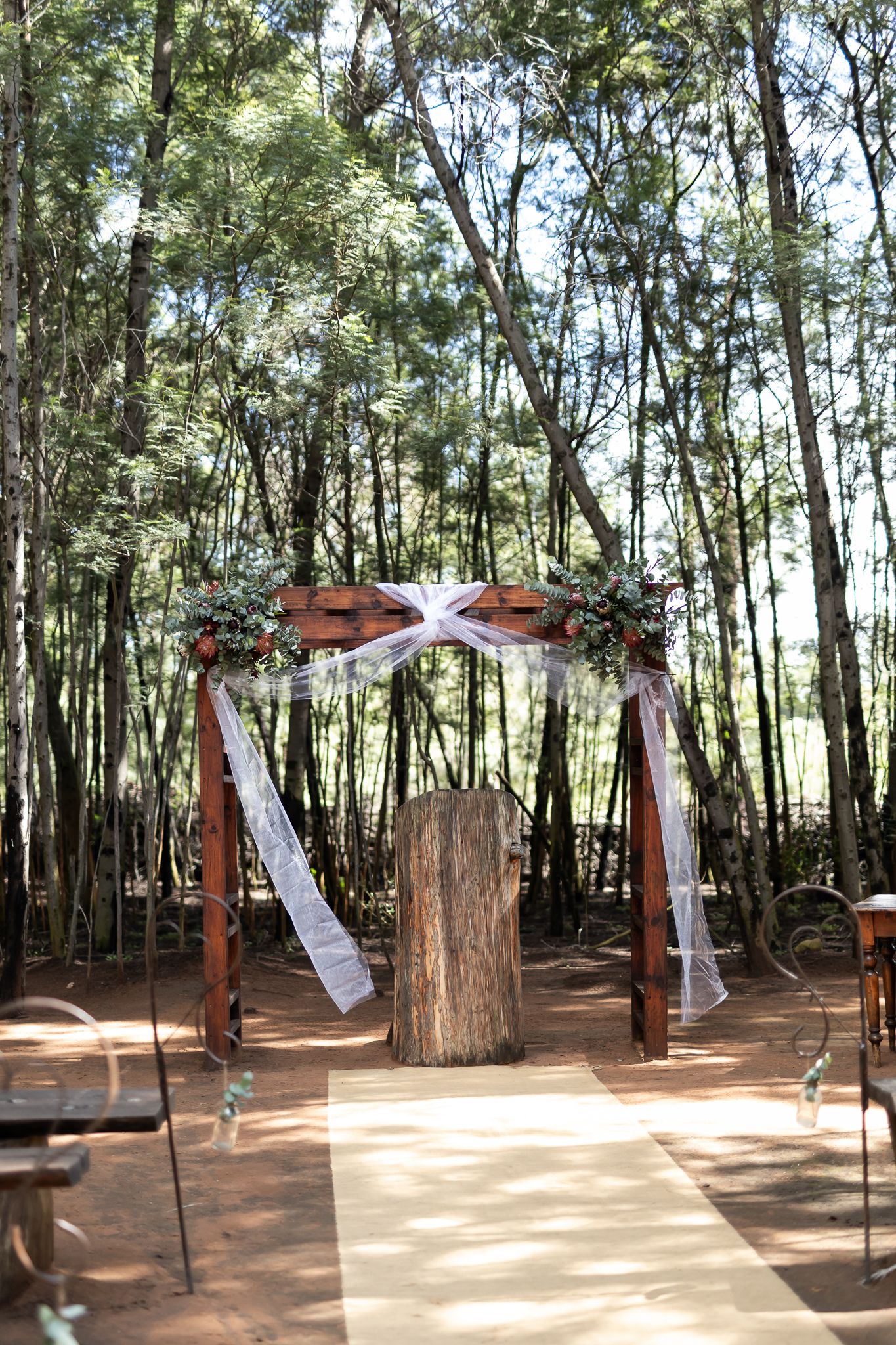 Rustic wooden wedding arch with white fabric and greenery in a forest setting.