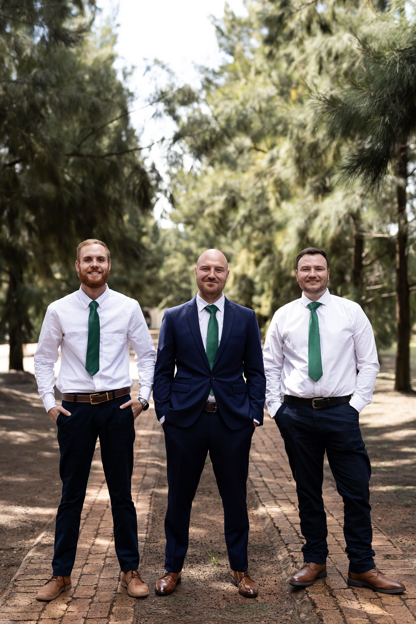 Three men in formal wear with matching green ties standing confidently on brick pathway.