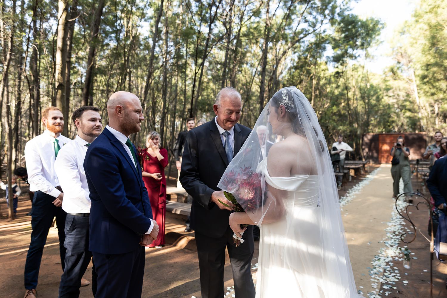 An intimate outdoor wedding ceremony taking place among tall trees with guests looking on.