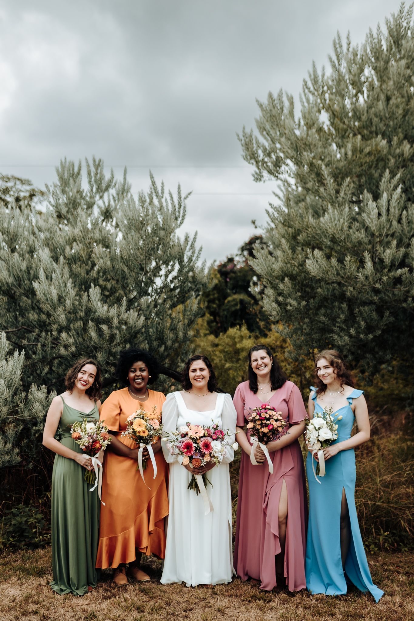 Bridal party in jewel-toned dresses pose together against evergreen trees.