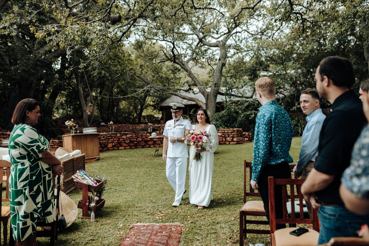 Outdoor garden wedding ceremony with guests seated on wooden chairs near stone walls.