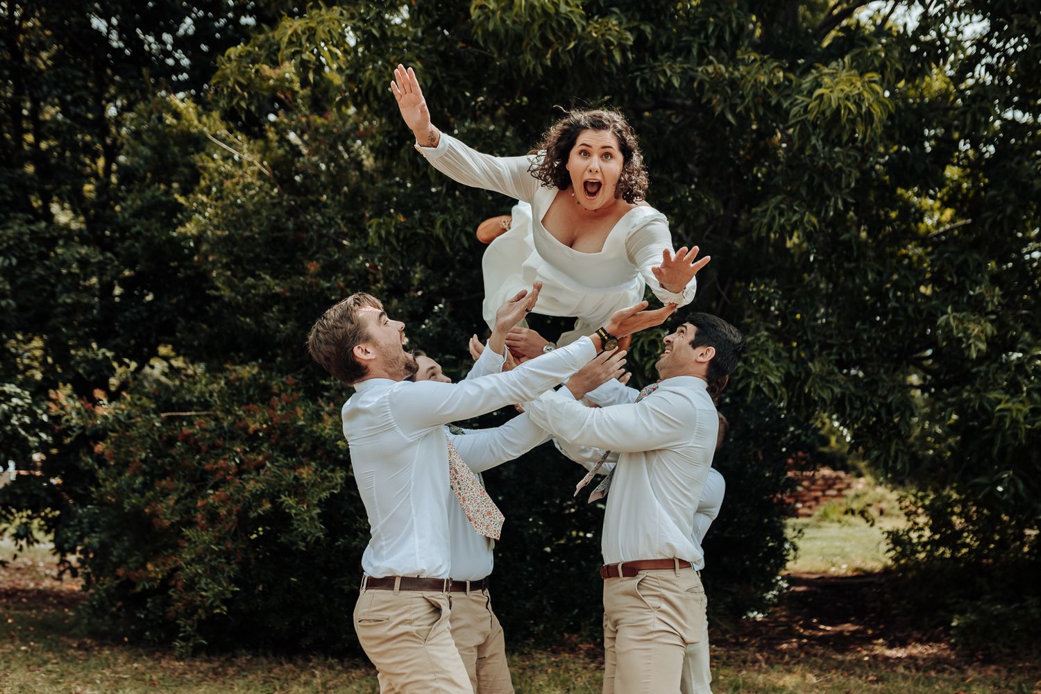 Playful wedding photo of people lifting someone in white dress outdoors.