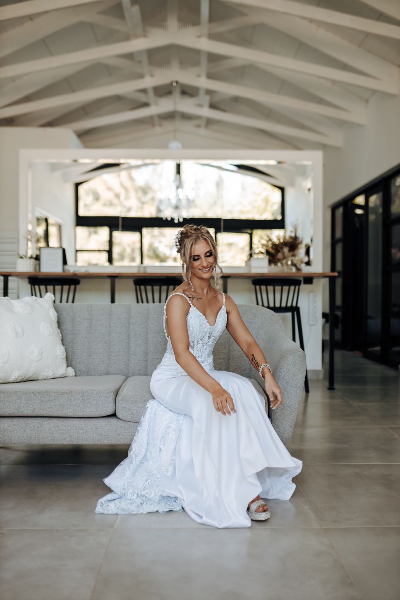 Person in white lace wedding gown poses elegantly on gray sofa in bright modern room.