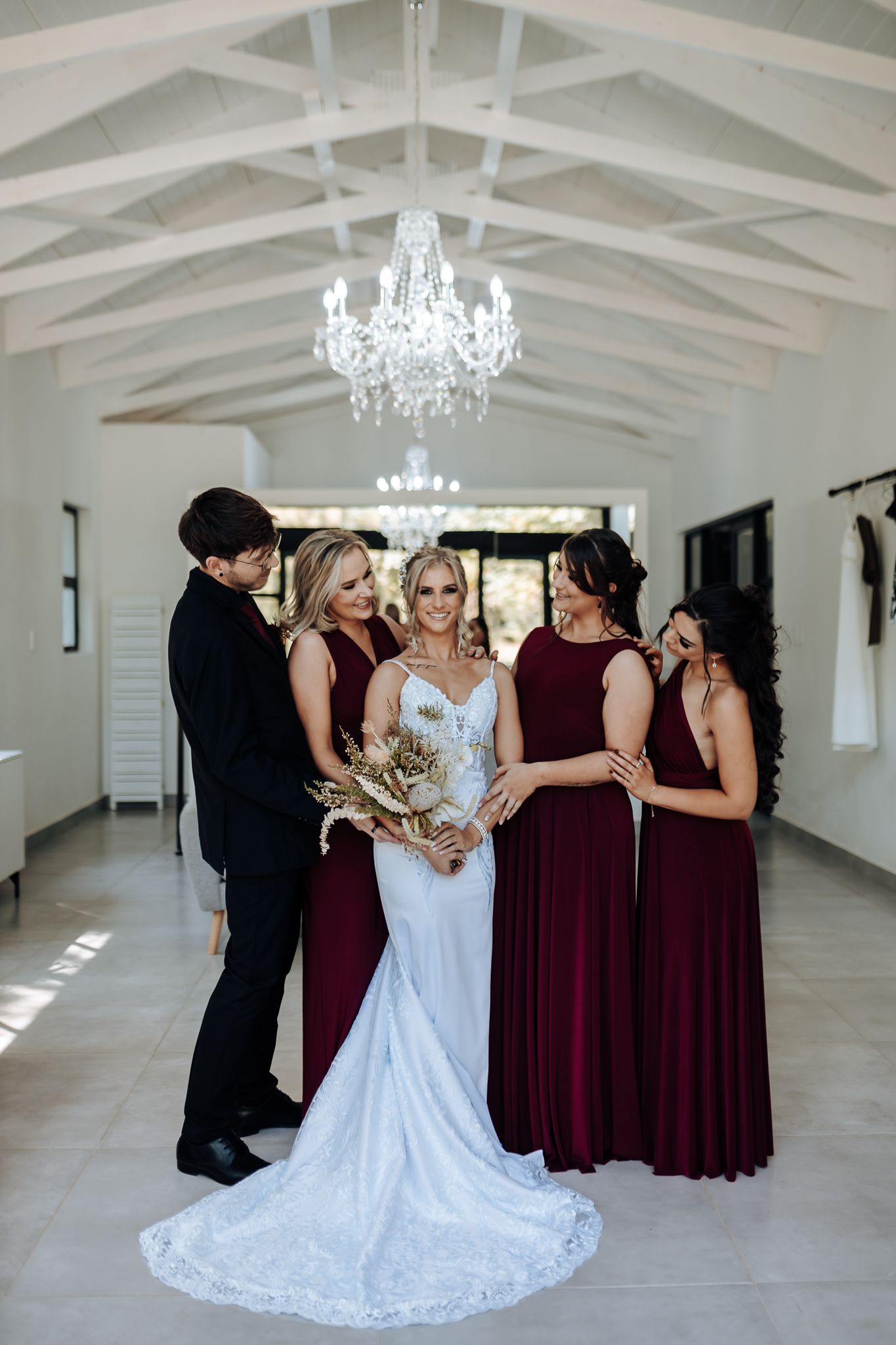 Wedding party in burgundy attire surrounds bride in fitted lace gown under crystal chandelier.