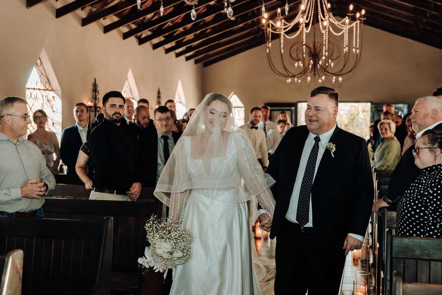 Wedding ceremony in progress inside rustic chapel with elegant chandelier and wooden beam details.