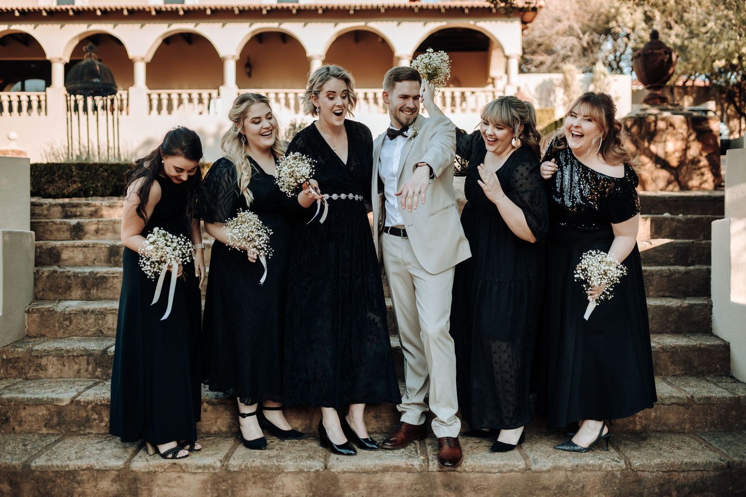 Wedding party in black dresses and suits poses on stone steps under arches at dusk.